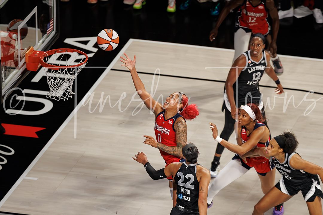 WNBA player in red uniform attempts a layup while defenders contest the shot near the basket.