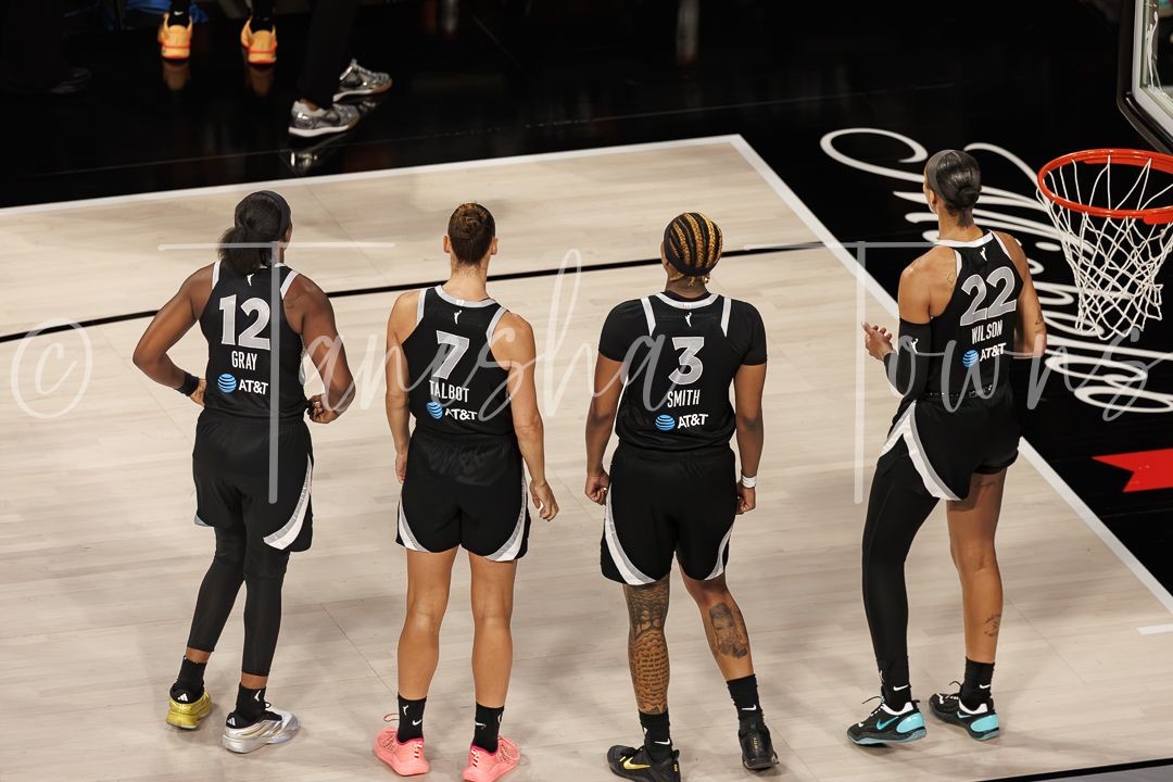 Four WNBA players in black uniforms stand on the court during a professional basketball game.