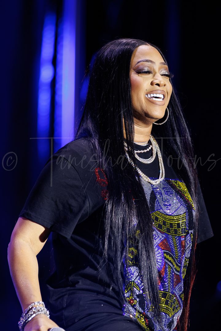 Female singer performing on stage with long black hair, wearing black graphic tee and layered pearl necklaces.