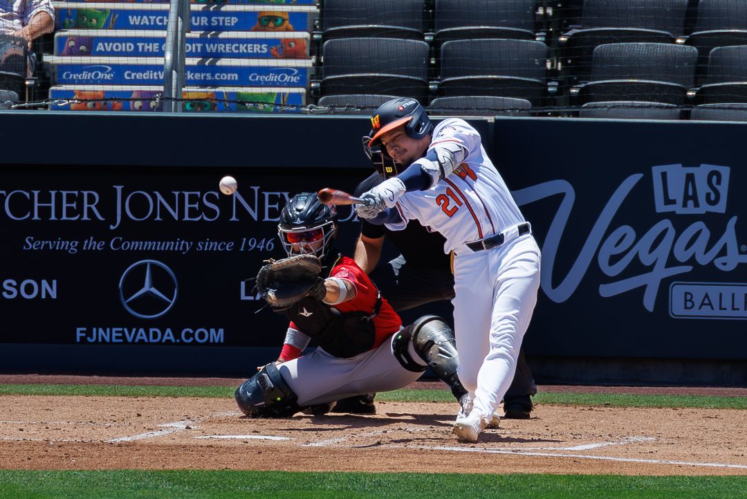 Baseball batter swings at pitch during Las Vegas Aviators game, catcher crouches behind home plate in sunny outdoor stadium.