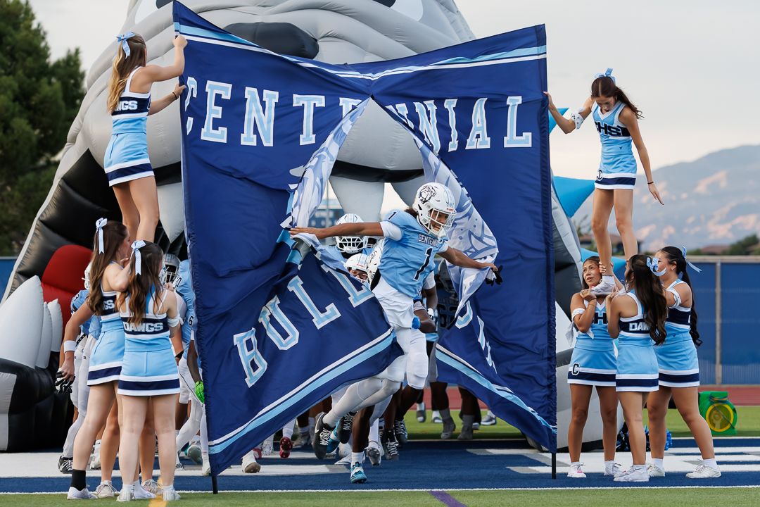 Centennial Bulldogs football player bursts through banner as cheerleaders perform stunts during game entrance.