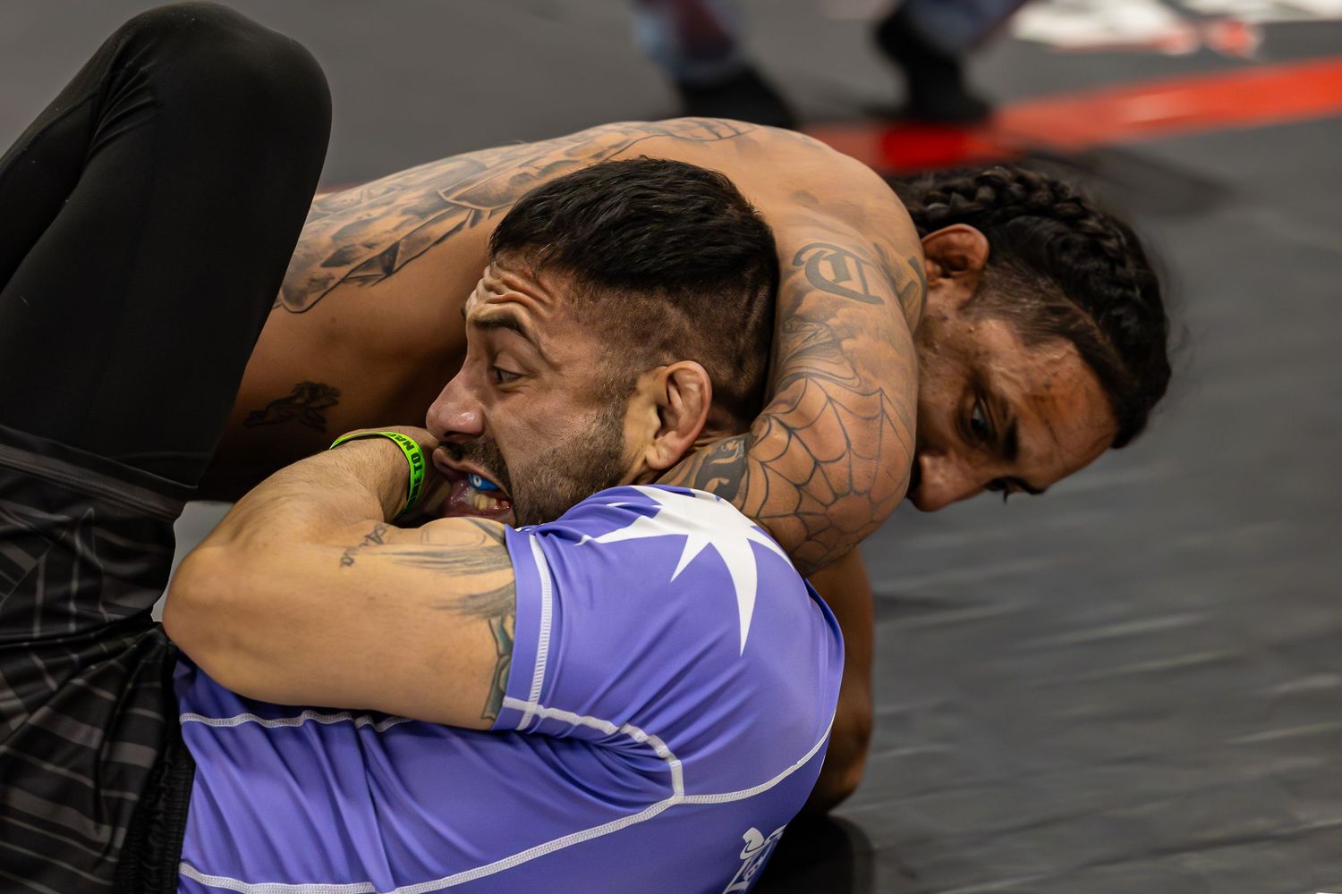 Two tattooed men grappling on a mat during a BJJ or MMA competition, one in a purple rash guard.