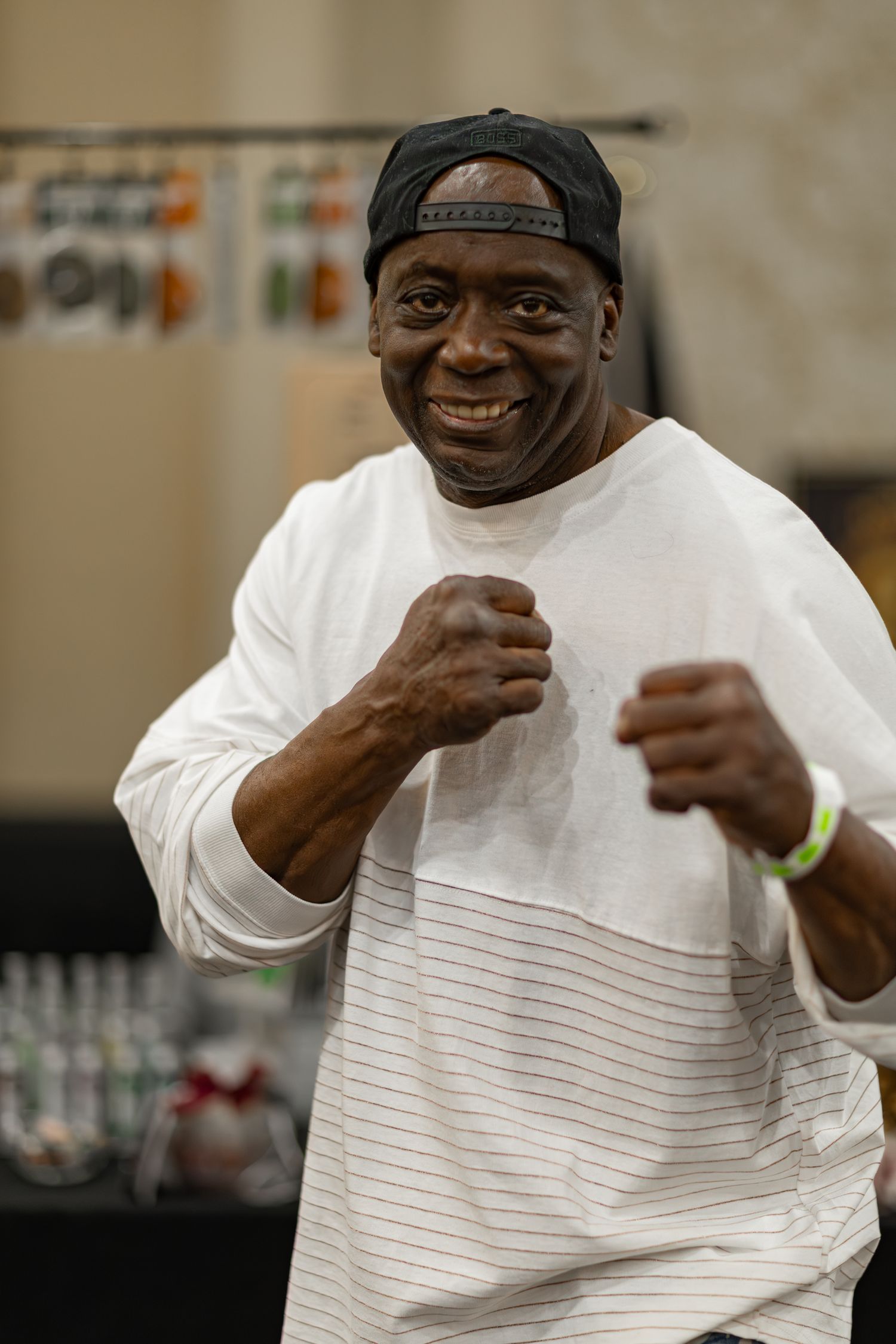 Smiling man in white shirt and black cap poses in boxing stance at indoor event.