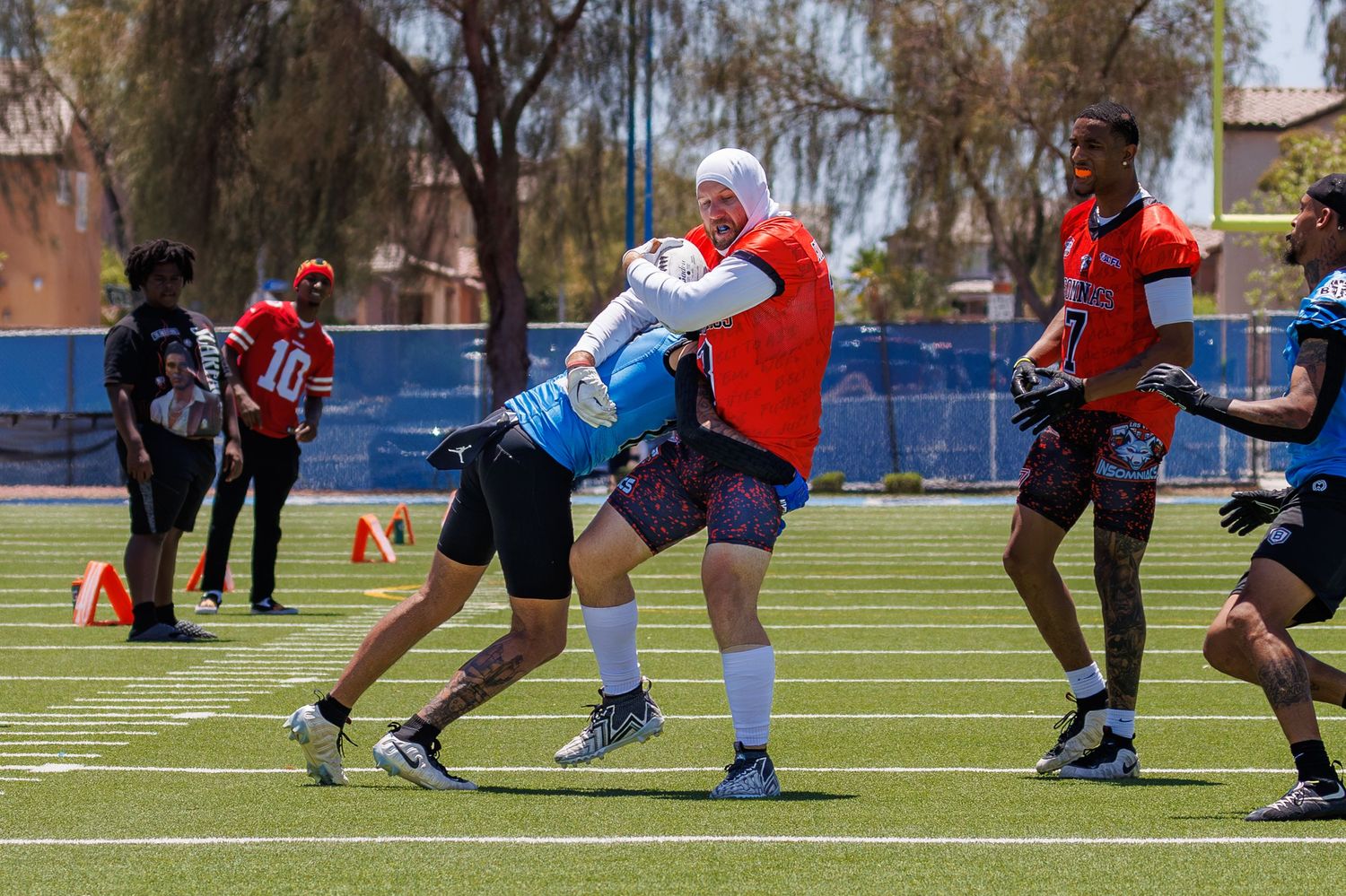 Football players collide during an outdoor practice drill on a blue turf field on a sunny day.