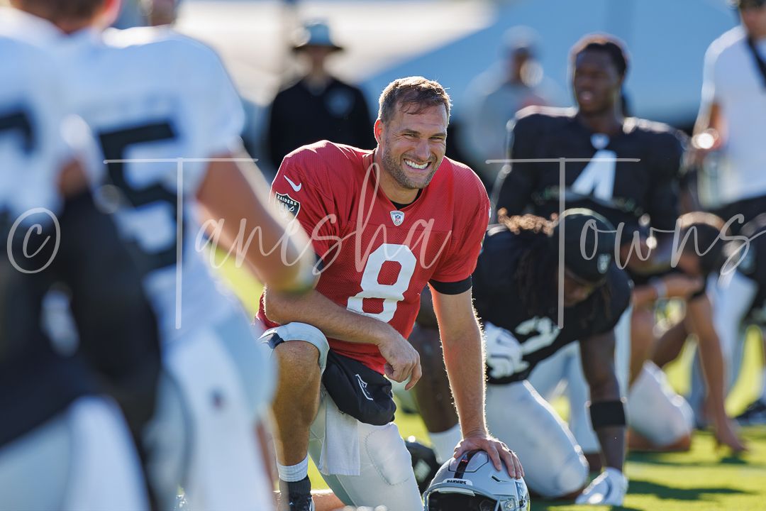 Smiling NFL player in red jersey number 9 kneeling on football practice field during Raiders training camp.