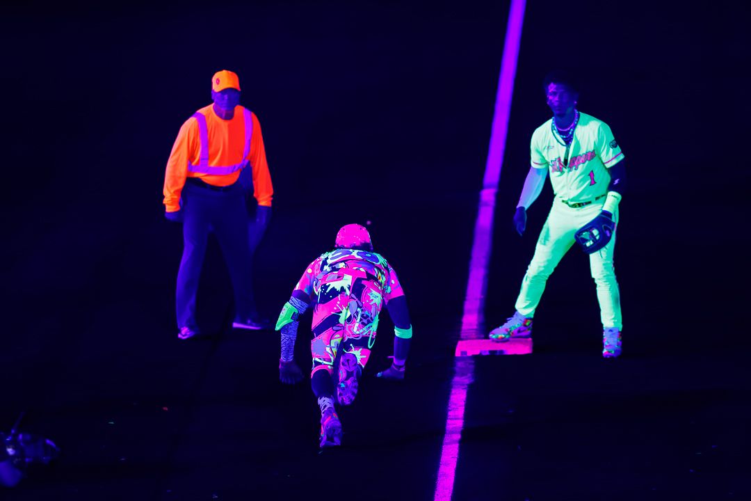 Three players glow under black light during a neon-lit baseball game on a dark field.