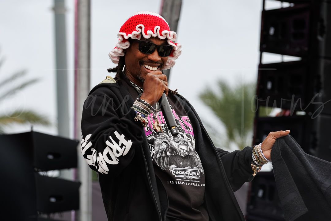 Male rapper performing on outdoor stage wearing red knit hat, sunglasses, black jacket, and gold chains.