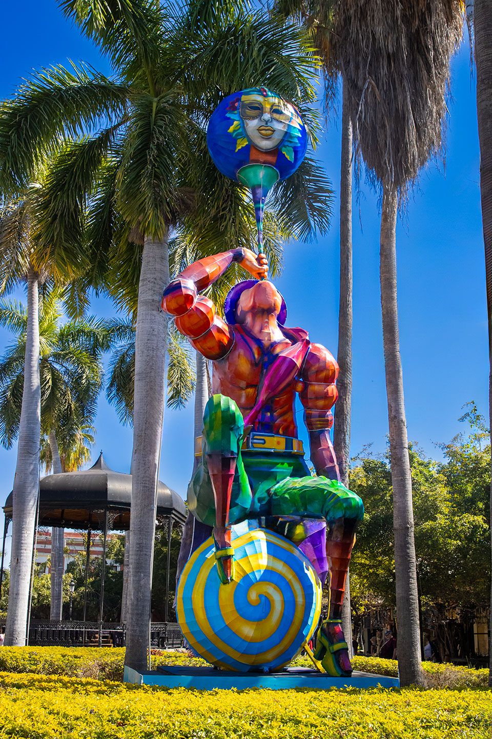Colorful whimsical sculpture of figures riding snails displayed in a tropical garden with palm trees.