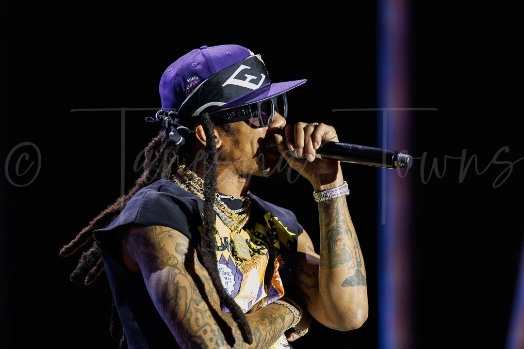 Tattooed rapper with dreadlocks and purple cap performing live on stage holding microphone.