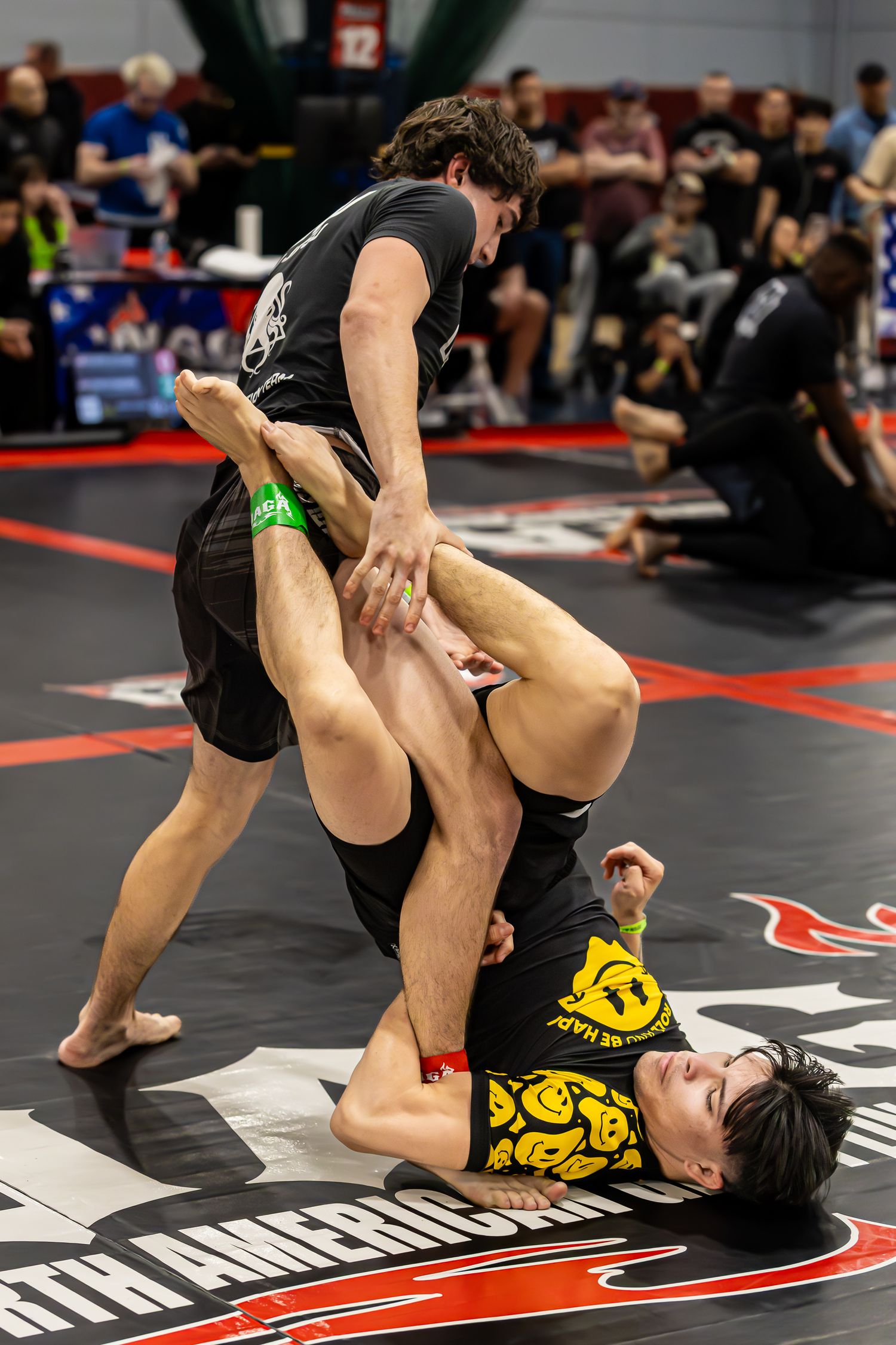 Two competitors grapple on the mat during a no-gi jiu-jitsu tournament, one applying a leg lock submission.