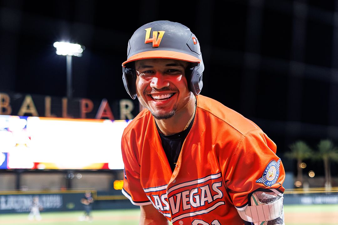 Smiling Las Vegas baseball player in orange uniform and LV helmet poses on field at night.