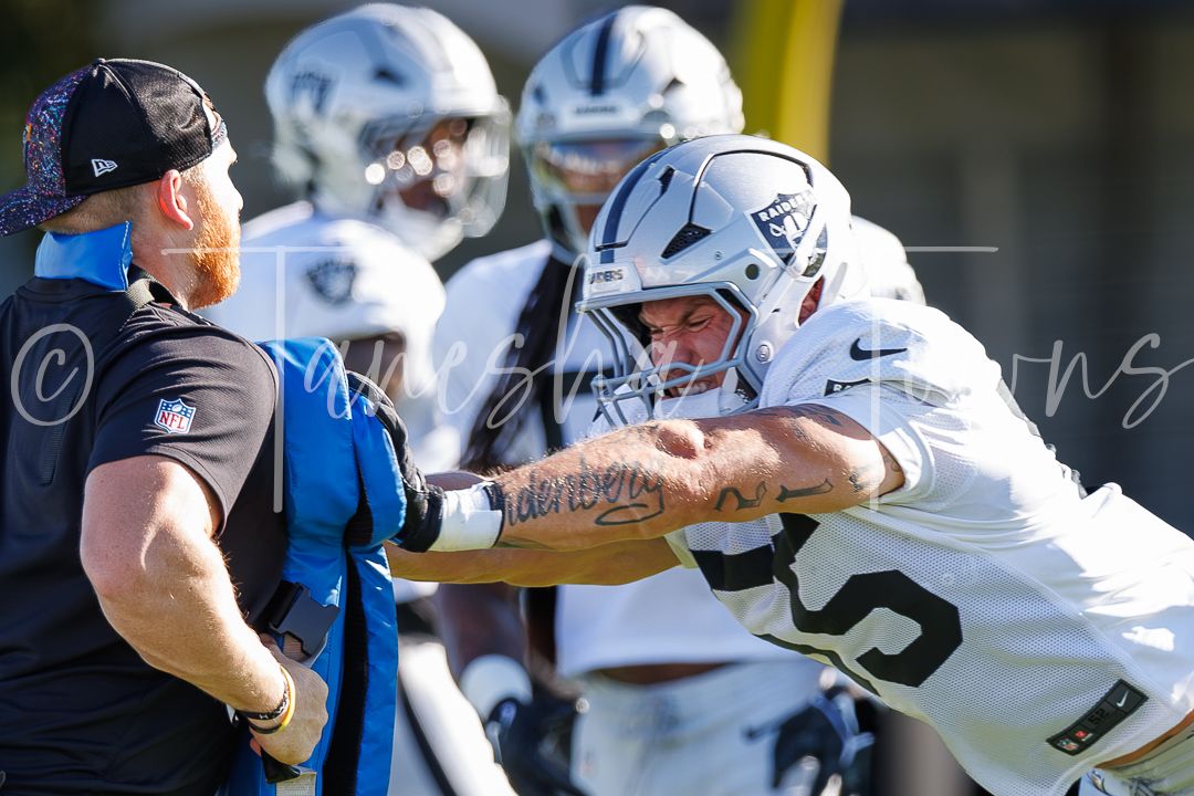 NFL players practice blocking drills in white uniforms during Raiders training camp on a sunny day.