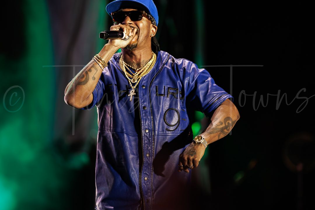 Male rapper performing on stage, wearing blue jersey and gold chains, singing into microphone under green stage lights.