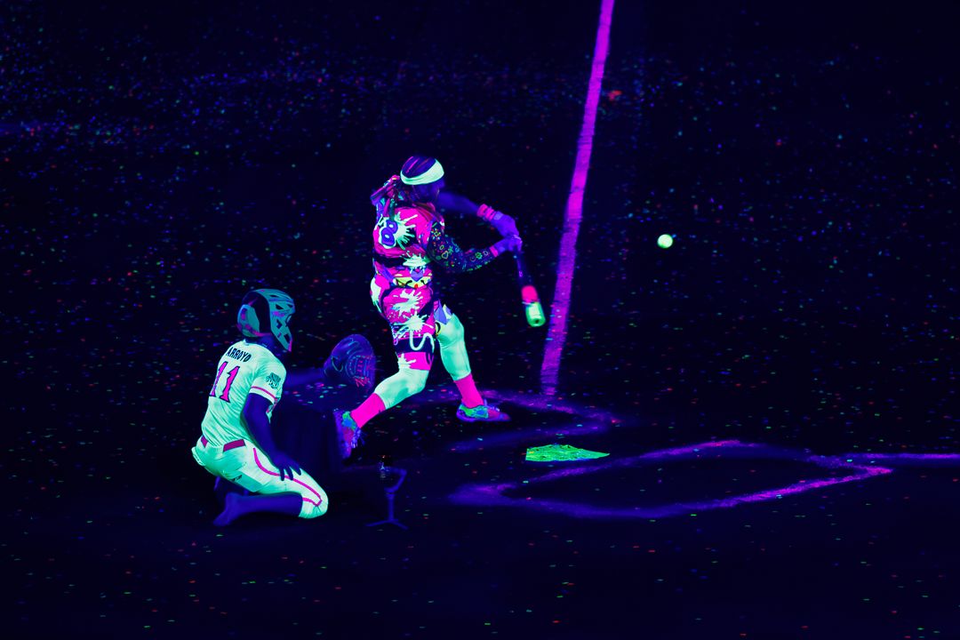 Players perform under black lights in neon glowing uniforms during a dramatic nighttime baseball game.