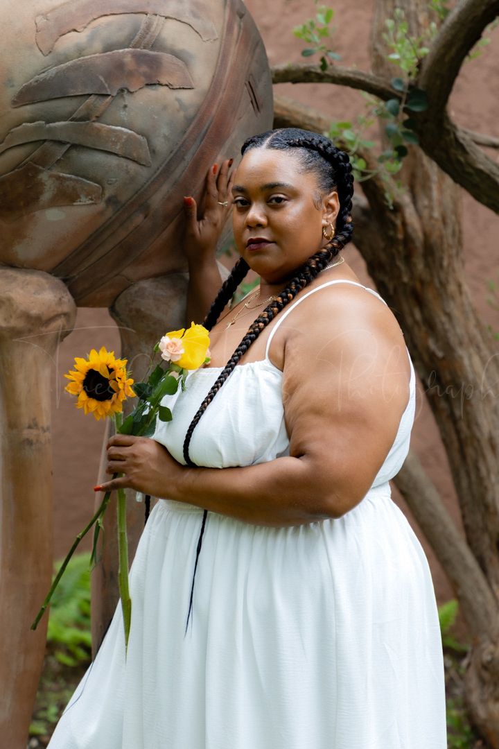 A person in a white dress holds sunflowers while posing against a rustic wall backdrop.