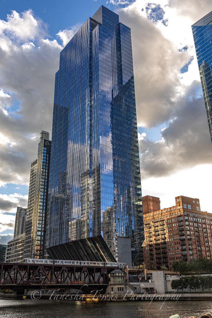 Blue glass skyscraper towers over Chicago River with dramatic cloudy sky and historic bridge in view.