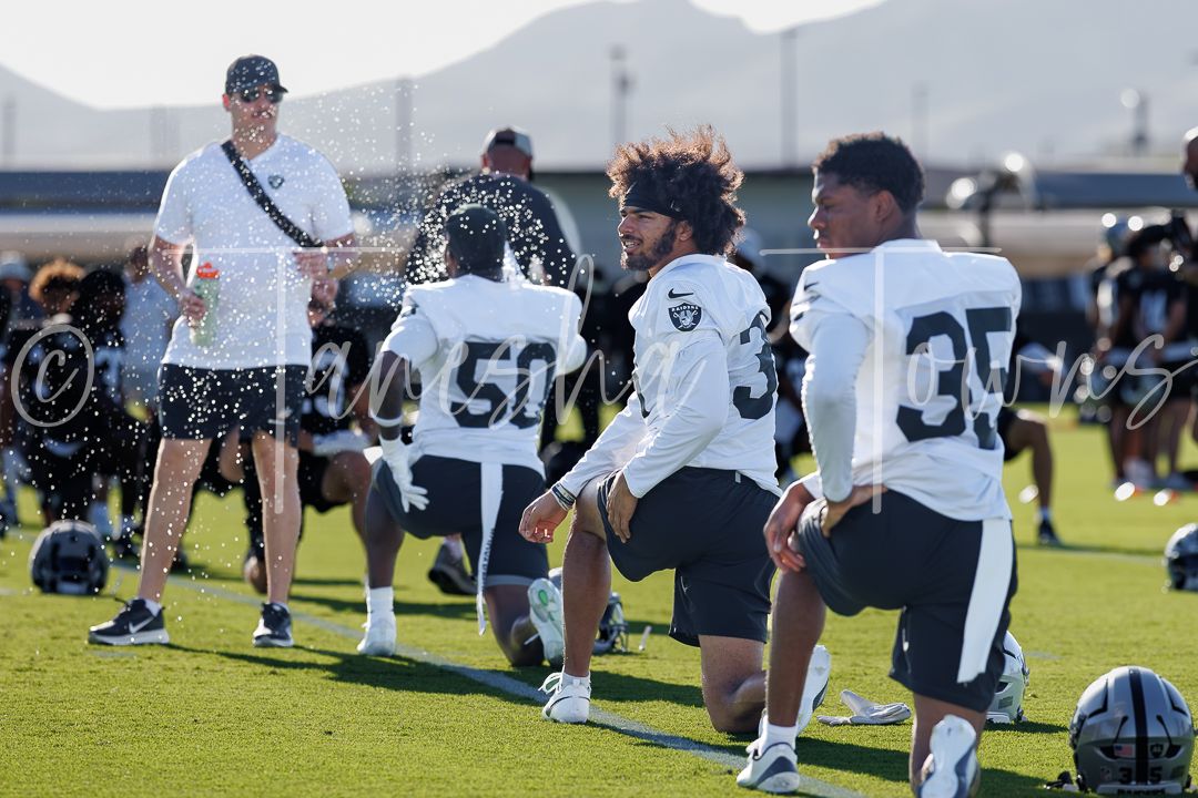 Raiders players in black jerseys cool off with water during an NFL training camp practice session outdoors.