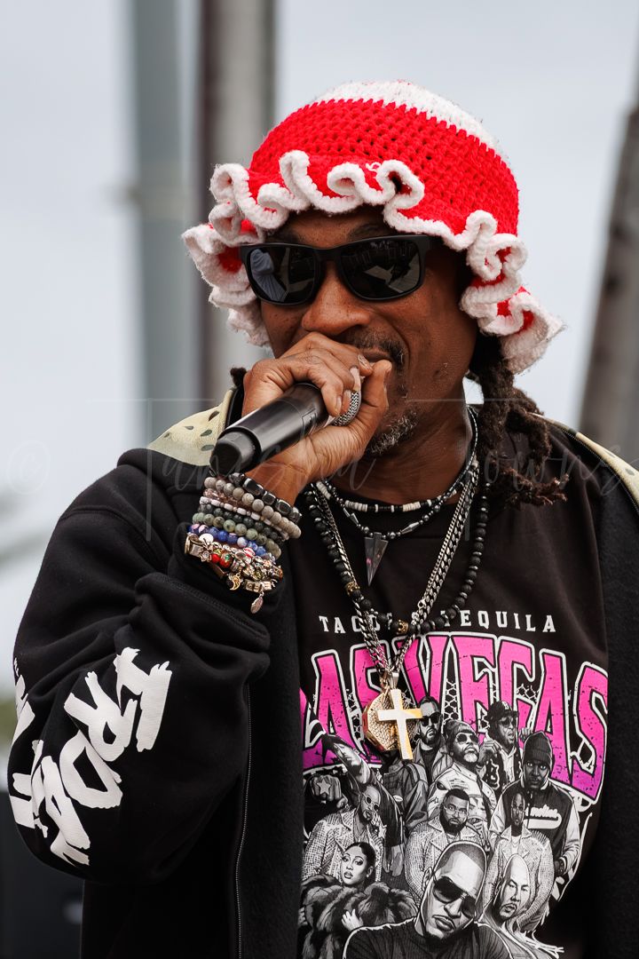 Performer in red crochet hat, sunglasses, Las Vegas graphic tee, and chains sings into microphone on outdoor stage.