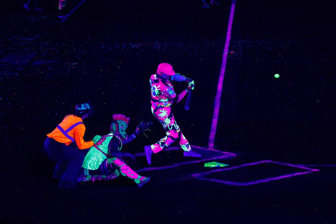 Athletes playing baseball under black light wearing neon UV-reactive uniforms and gear.