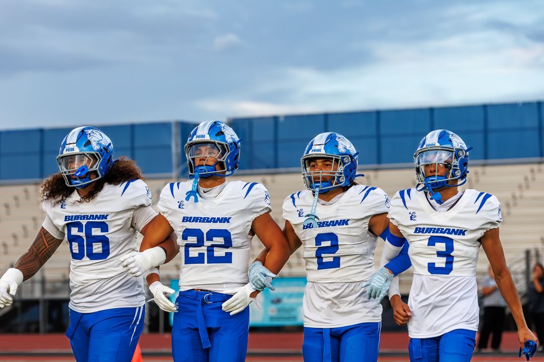 Four Burbank high school football players in blue and white uniforms walk together on the field.