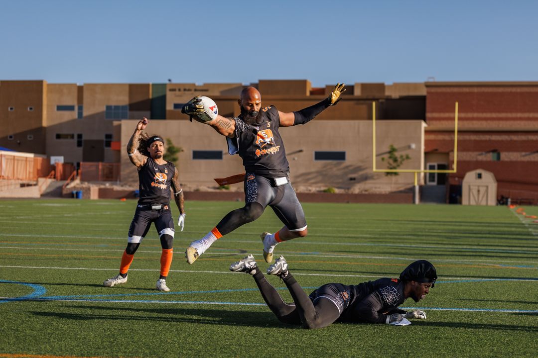 An athlete leaps to catch a football while a defender falls on a sunny turf field during practice.