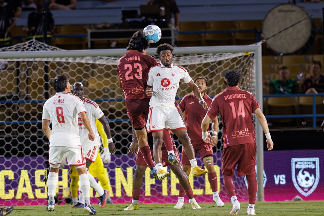 Soccer players compete for a header during a night match, with player #23 jumping above opponents on the field.