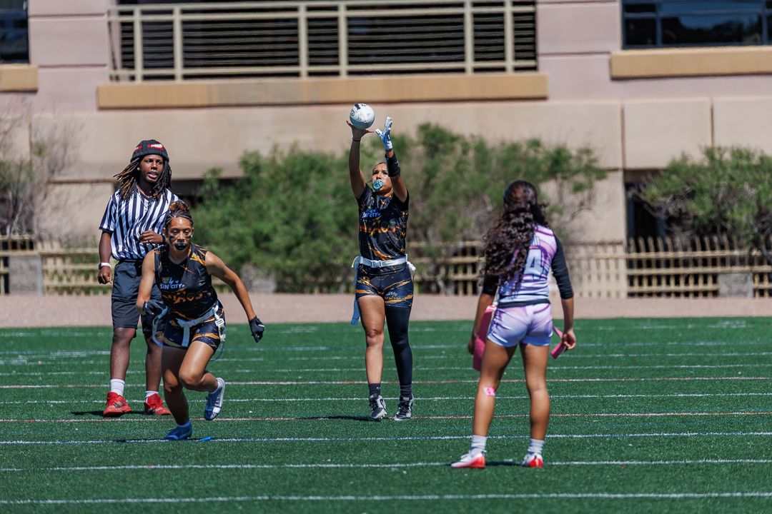 Women playing flag football on a green turf field with a referee watching during a sunny outdoor game.