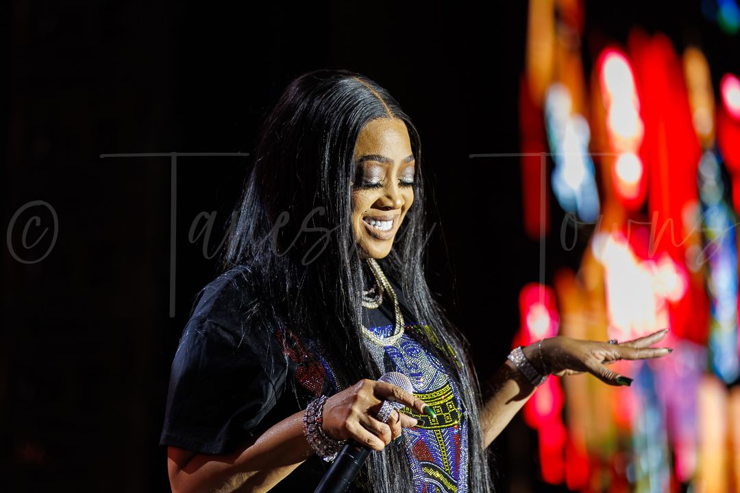 A woman with long black hair performs on stage, gesturing with her hands against a colorful backdrop.