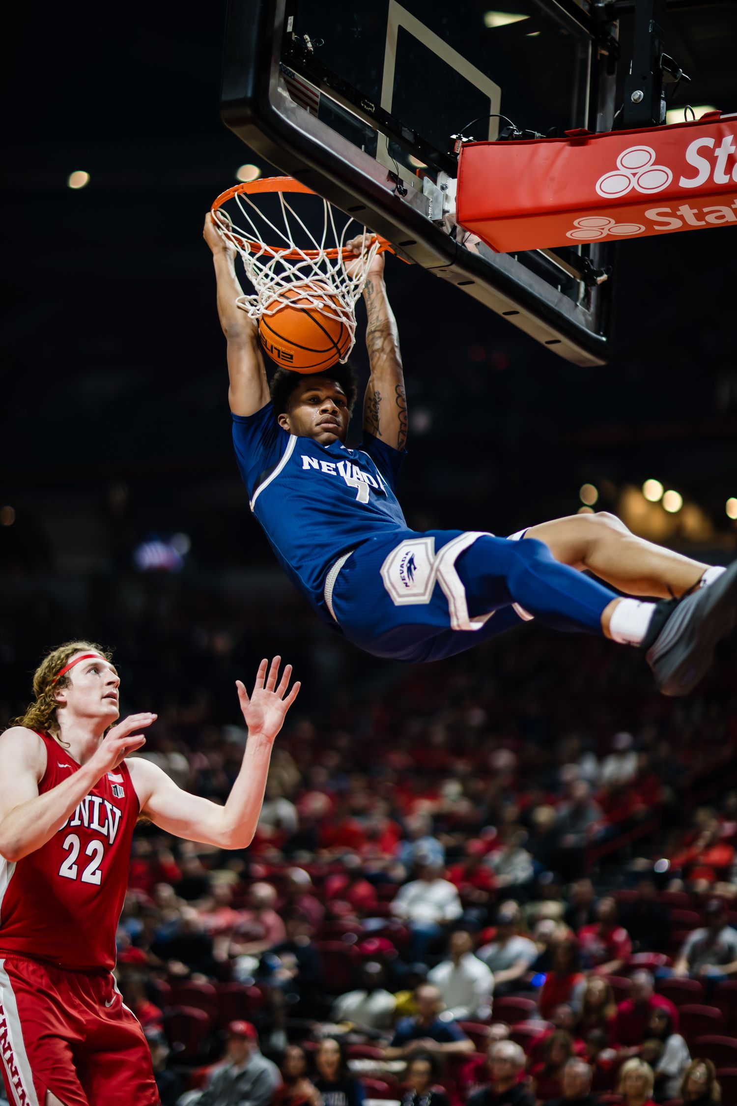 Nevada basketball player dunks over UNLV defender #22 in a packed arena during a college basketball game.