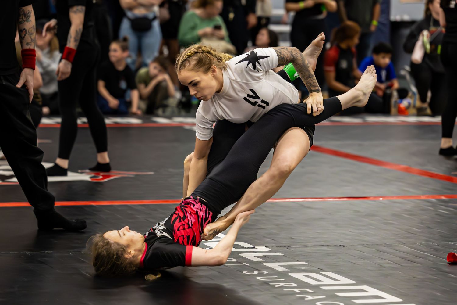 Two women competing in a grappling martial arts match on a black mat at an indoor tournament.