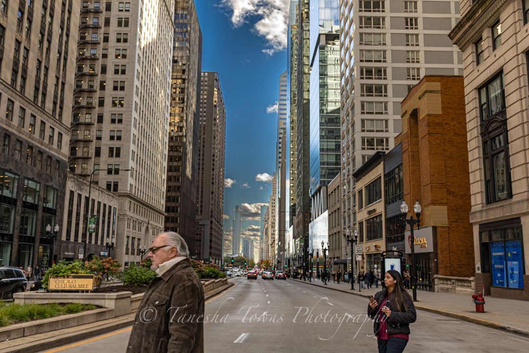 Wide-angle urban street view of downtown Chicago with tall skyscrapers and pedestrians on a sunny day.