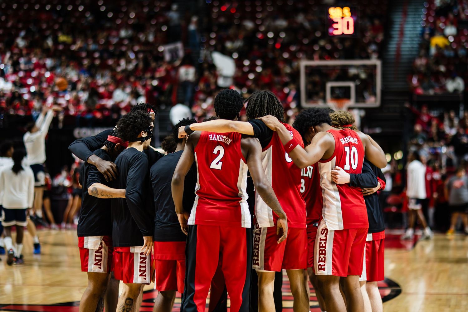 College basketball team huddles together on court during a game in a packed arena wearing red uniforms.