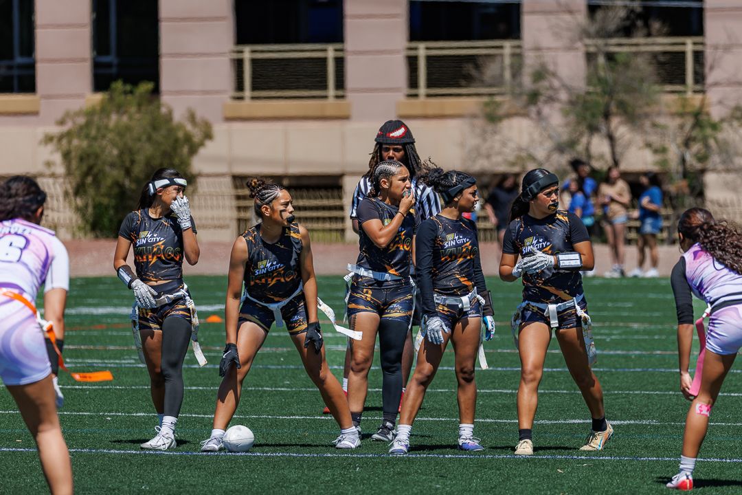 Women playing flag football on a green turf field during a sunny outdoor game.