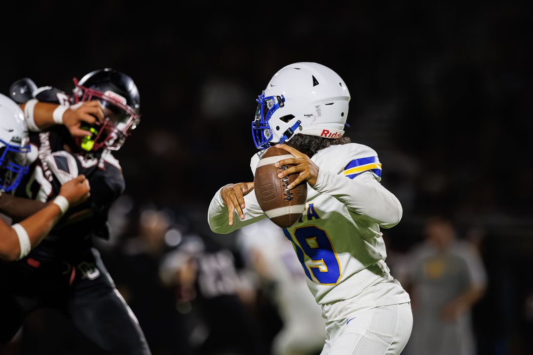 Football quarterback in white uniform drops back to pass during a night game under bright stadium lights.