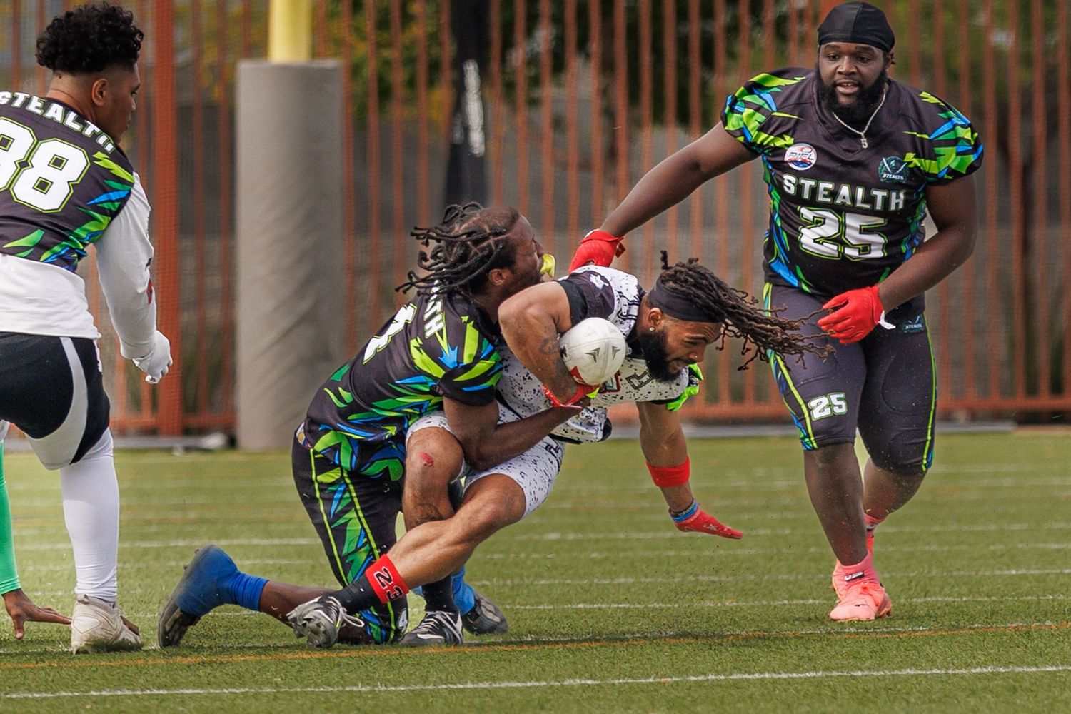 Flag football players in green Stealth uniforms compete during an intense tackle play on a turf field.