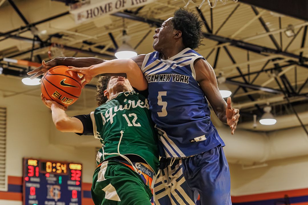Basketball player #12 in green drives to the basket against a defender in blue during an indoor game.