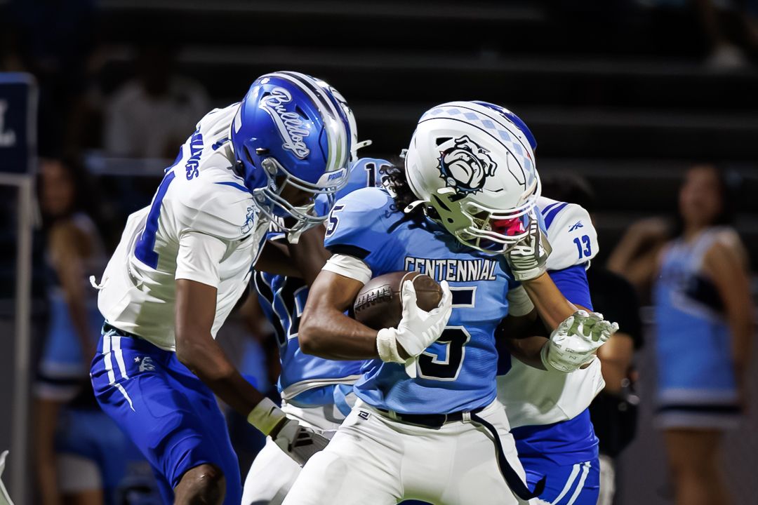 Football players in blue and white uniforms compete during a night game, with a Centennial player carrying the ball.