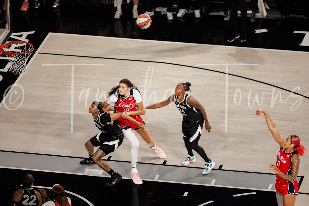 WNBA players compete for the ball during an intense basketball game, with players in black and red uniforms driving to the basket.