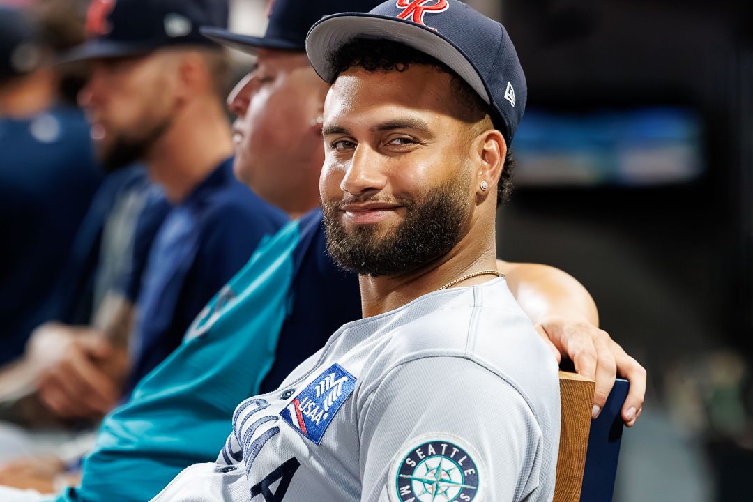 Smiling baseball player in Seattle Mariners dugout wearing gray uniform and navy cap during MLB game.