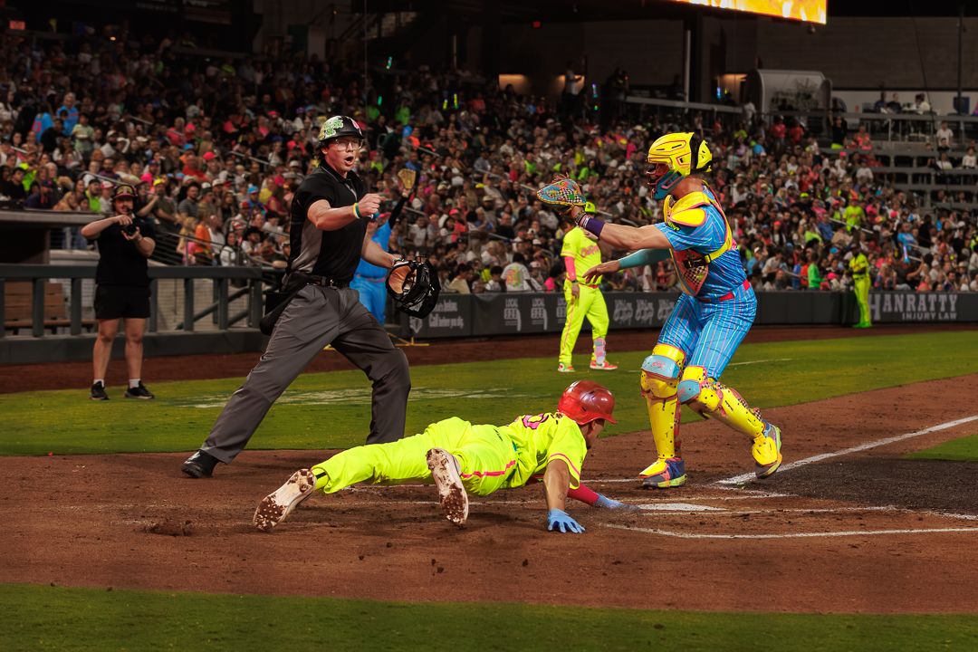 Baseball player slides into home plate as catcher attempts tag during a colorful night game with umpire watching.