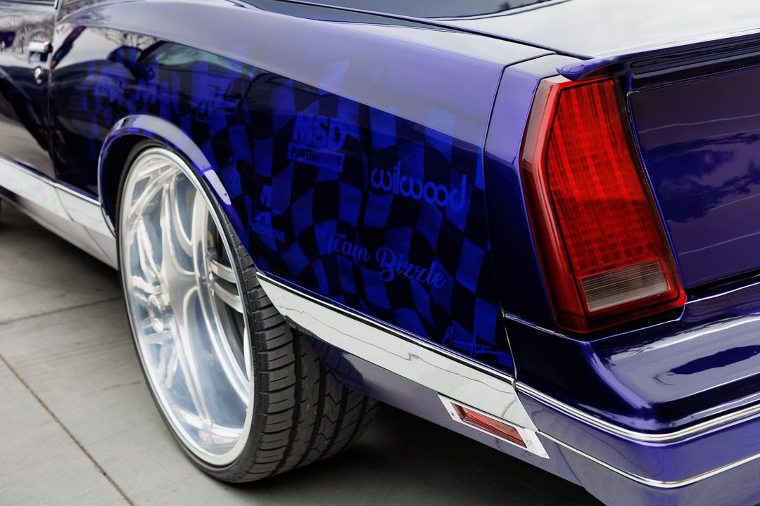 Custom purple checkered lowrider car with chrome wheels and red tail light detail.