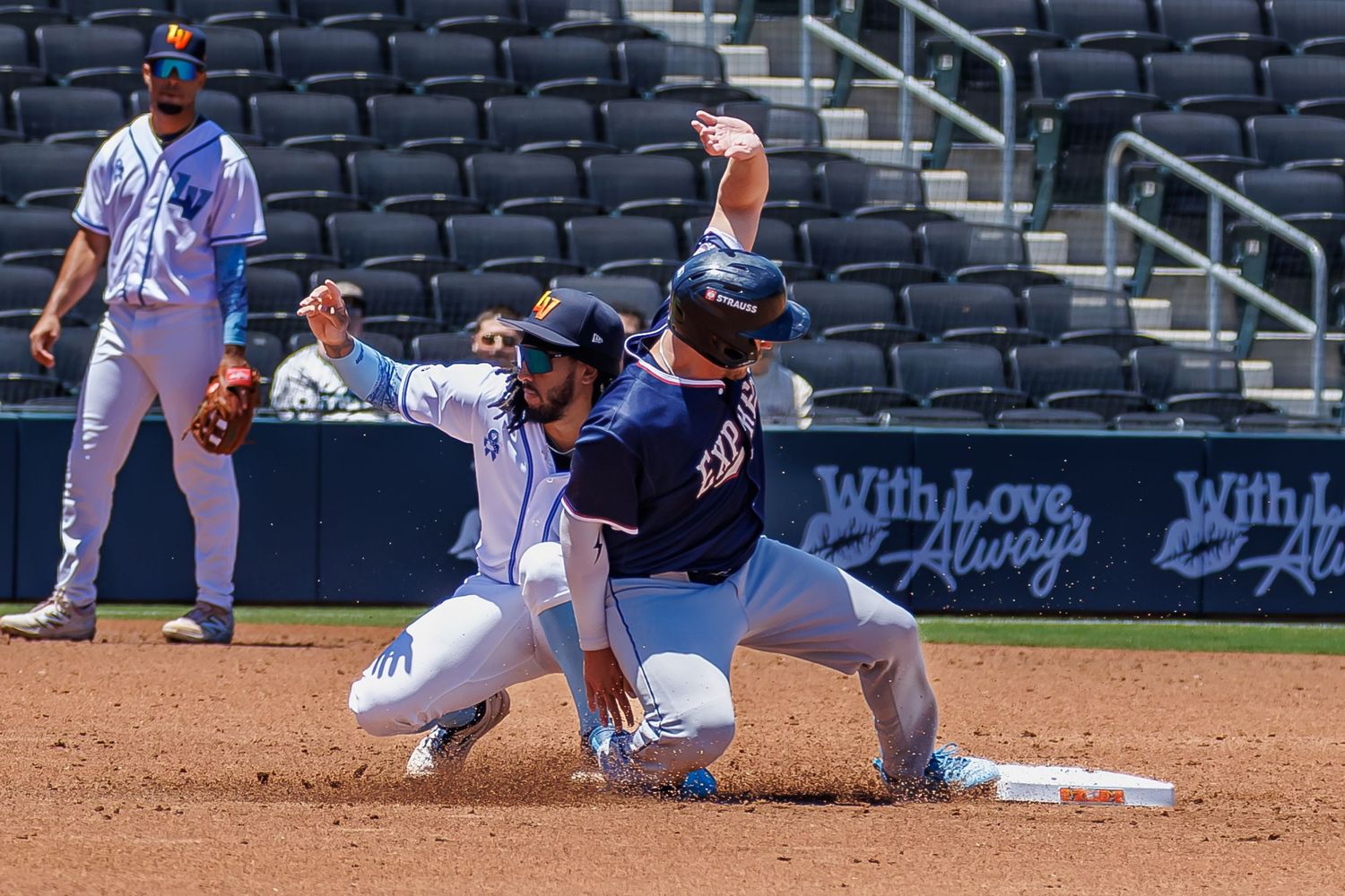 Baseball player sliding into base as fielder attempts tag during sunny day game at professional stadium.