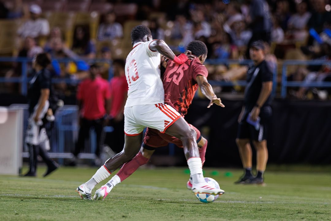Two soccer players battle for the ball during a night match, one in white and one in red uniforms.