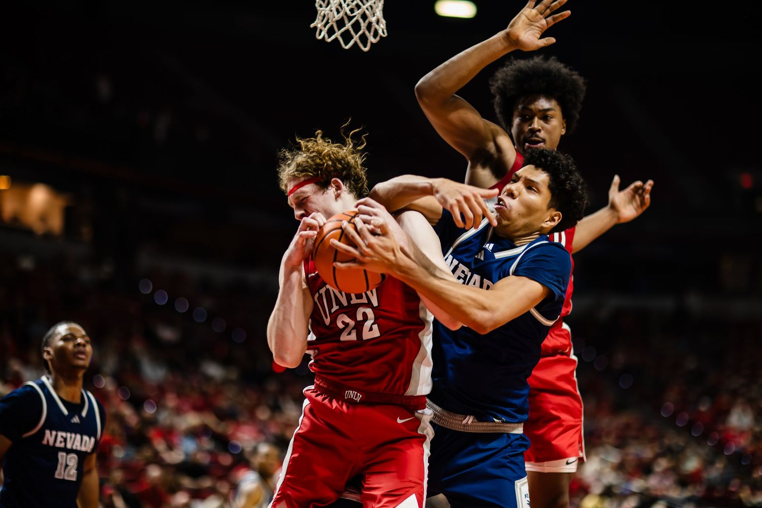 College basketball players battle for a rebound during an intense game, with players in red and navy uniforms competing.
