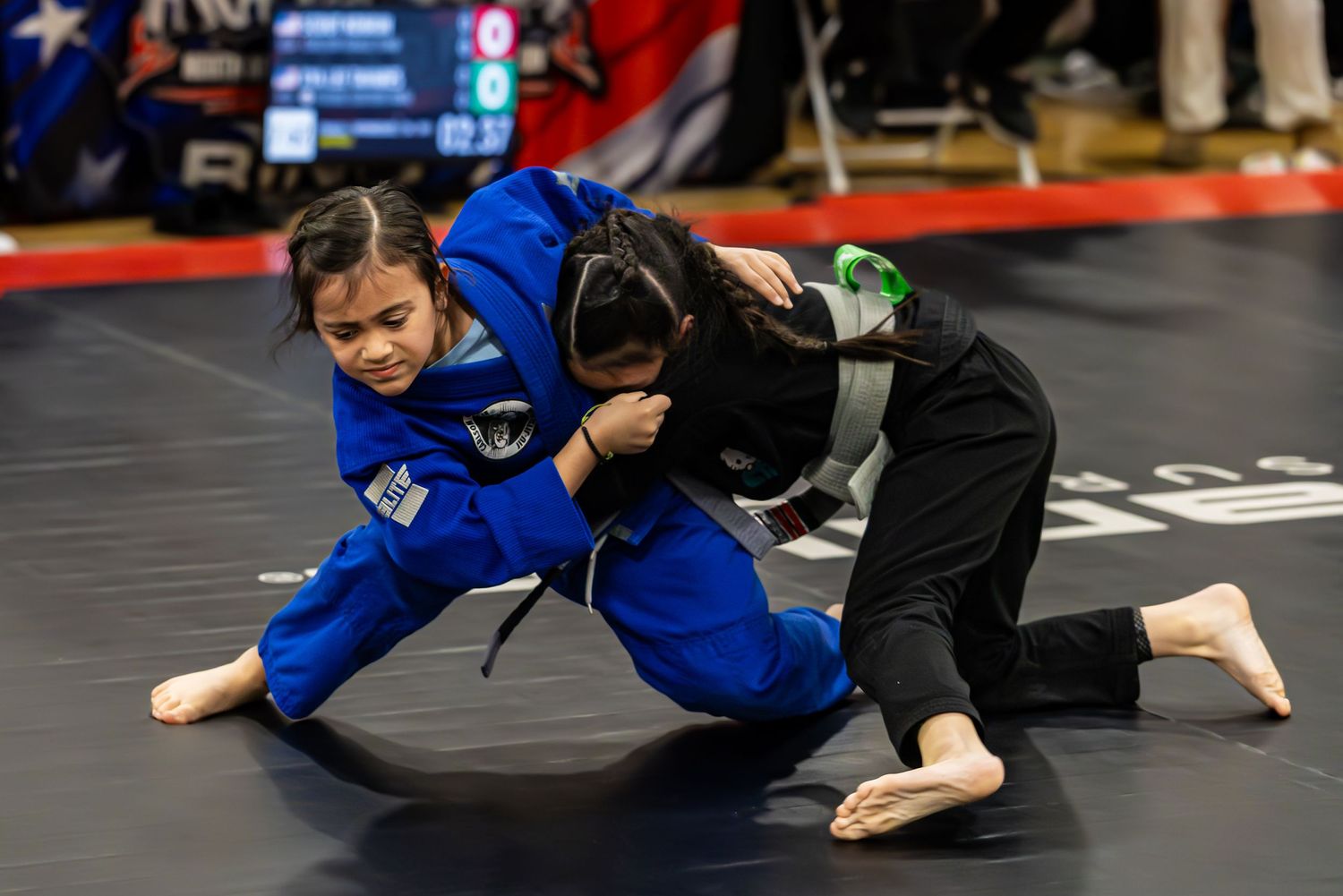Two young girls competing in a Brazilian jiu-jitsu match, one in blue gi and one in black gi, grappling on a mat.