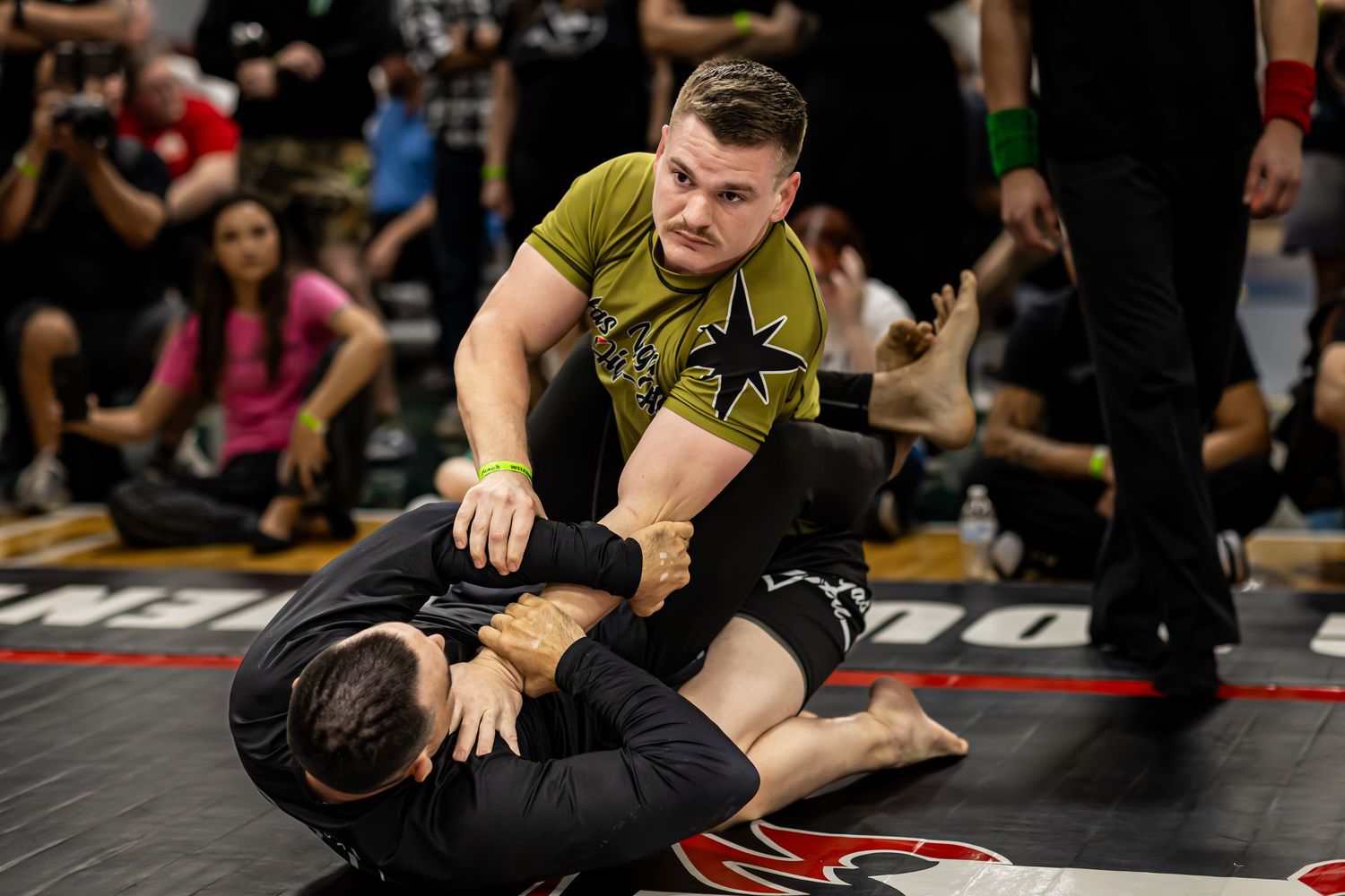 Two athletes compete in a grappling martial arts match on a mat at an indoor tournament.