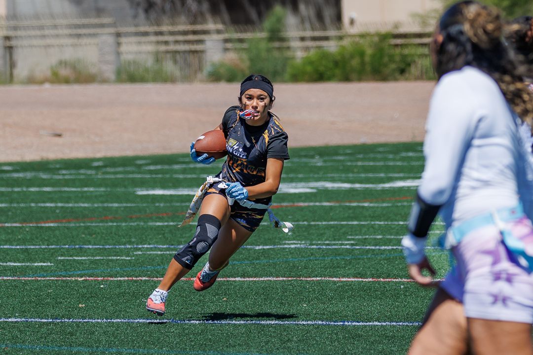Female flag football player runs with ball during outdoor game on turf field in athletic gear.