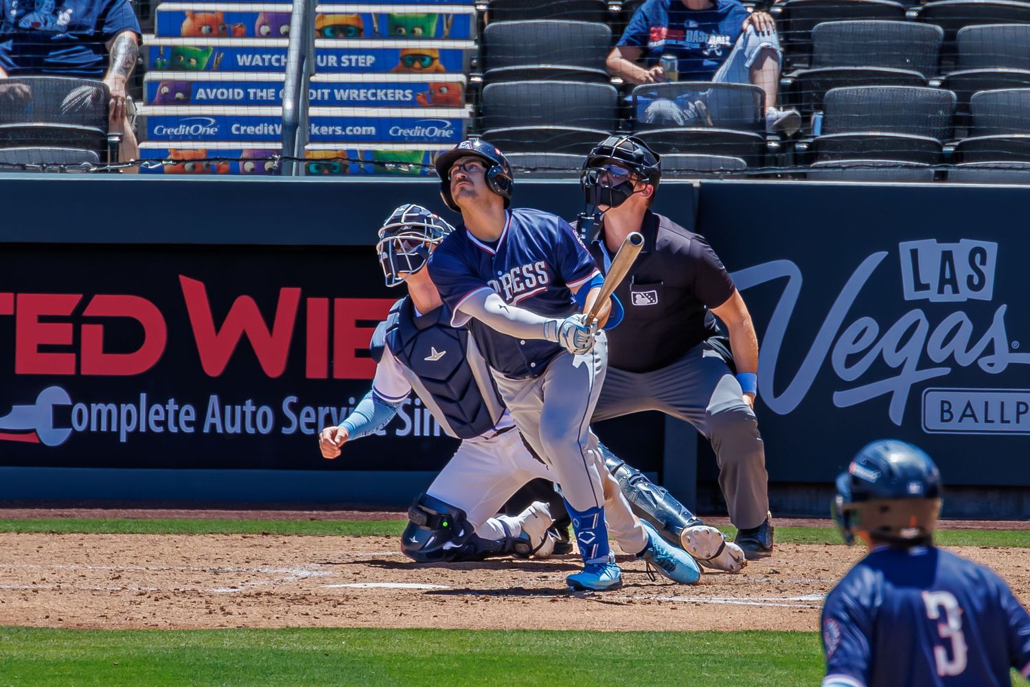 Baseball batter swings at pitch during Las Vegas Ballpark minor league game with catcher and umpire behind home plate.