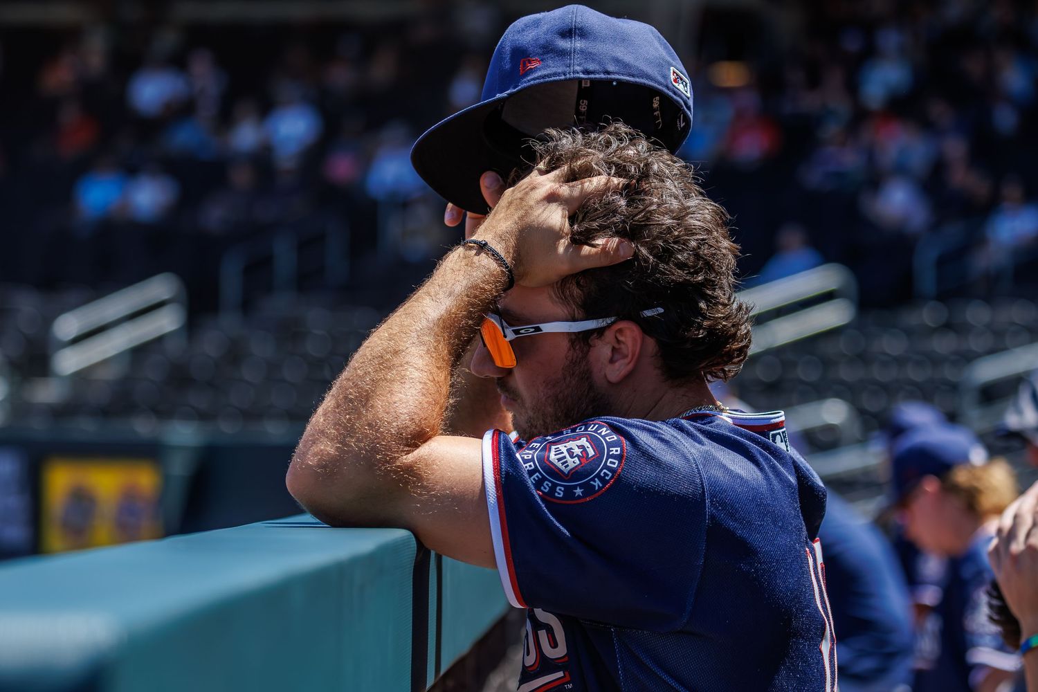 Baseball player in navy uniform and cap shields eyes from sun in stadium dugout on a bright day.