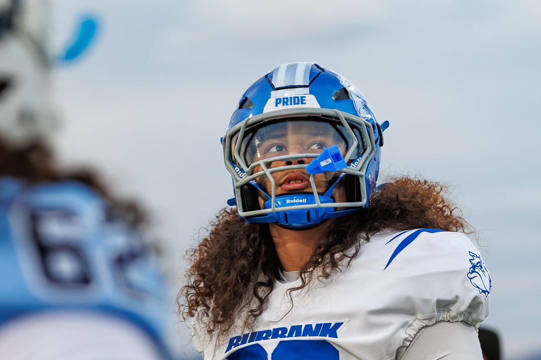 Football player in blue and white Burbank helmet with curly hair on the sideline during a game.